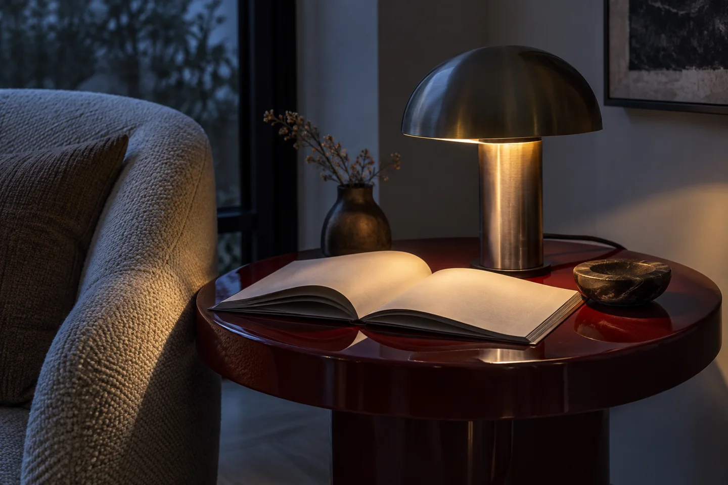 A sculptural metal reading lamp casts warm light over a book beside an upholstered chair.