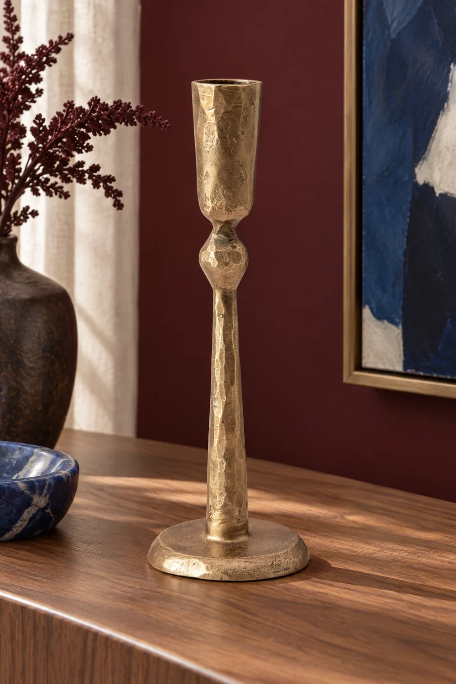 A single antique-brass-finish taper holder on a wood console beside framed art.