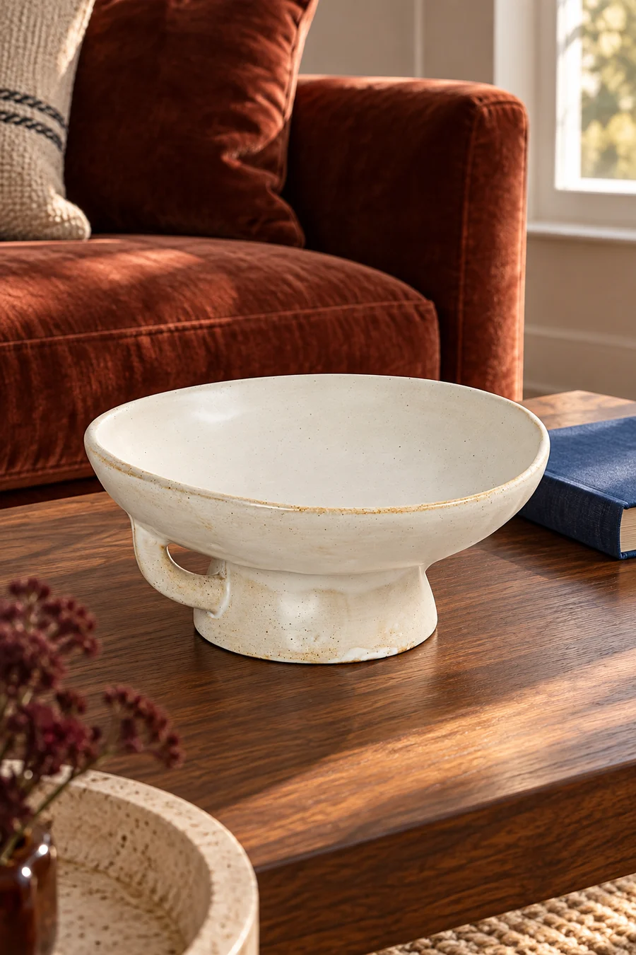 A cream pedestal bowl with a small loop handle on a walnut coffee table beside a rust sofa.